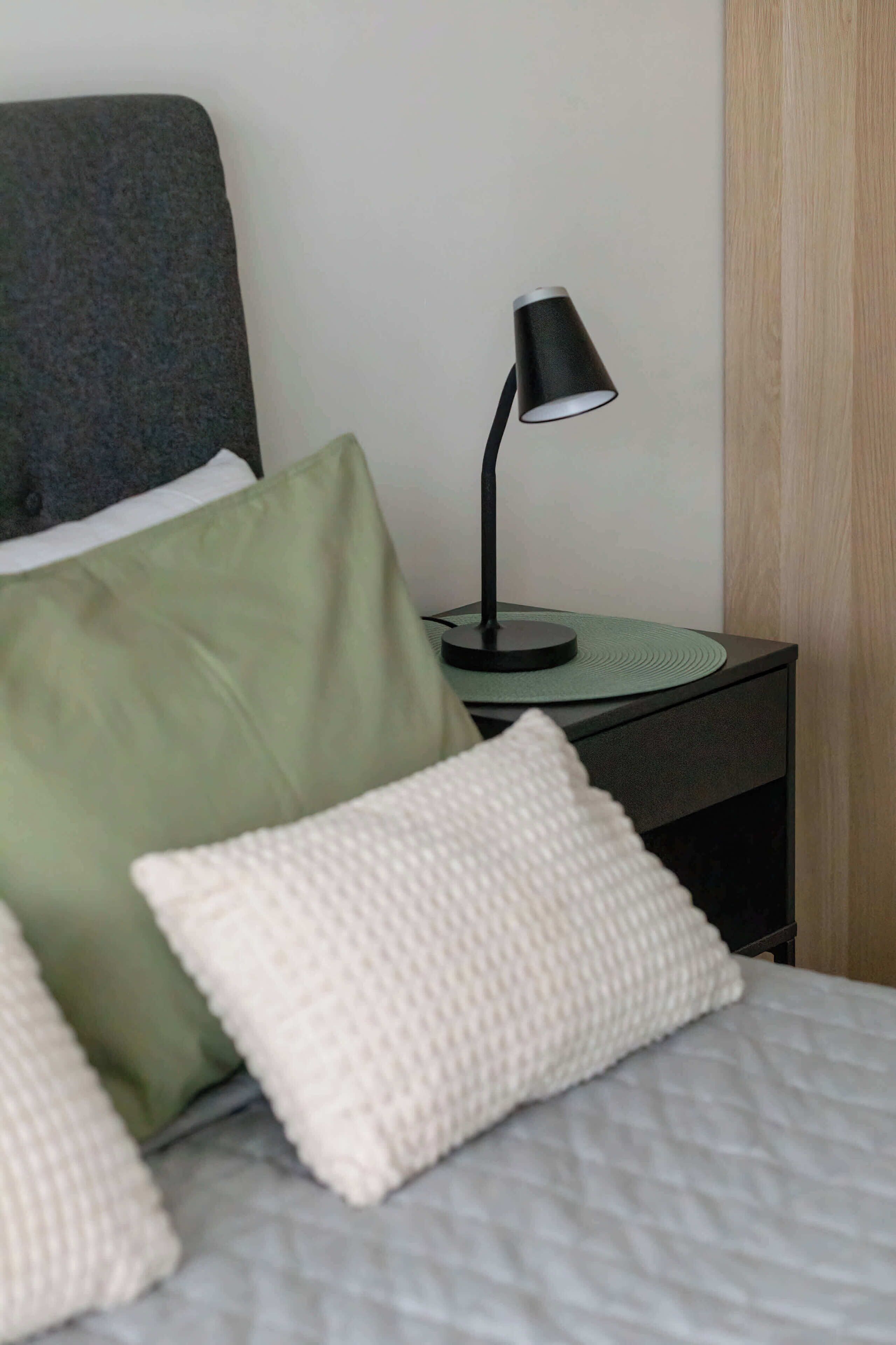 A neatly made bed with a grey quilt, a green pillow, and a white knitted pillow. A black desk lamp with a green base is on a black nightstand beside the bed.