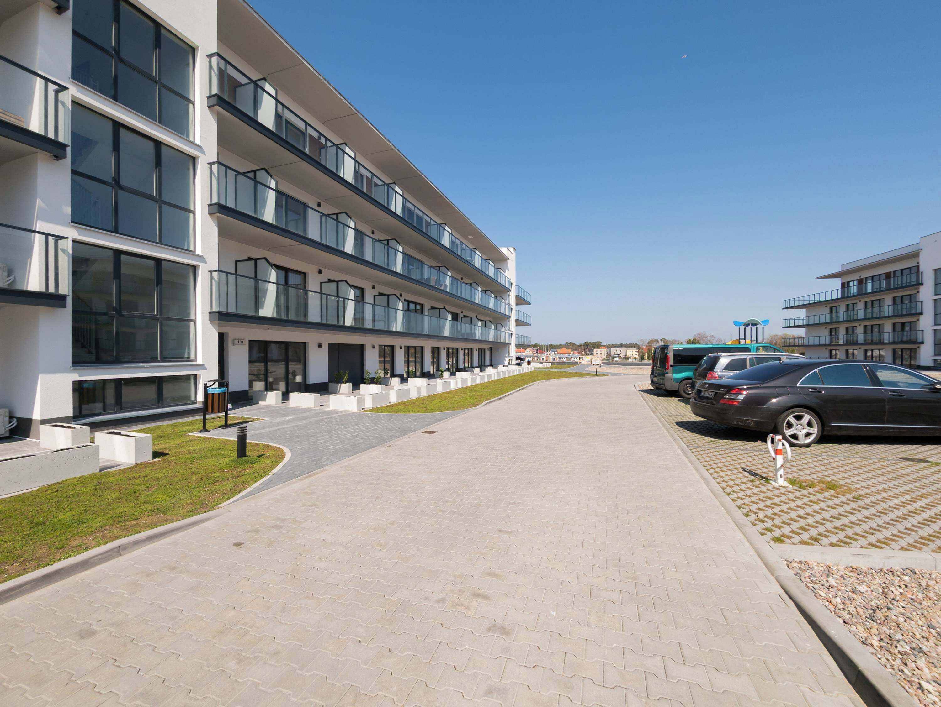Un bâtiment moderne à plusieurs étages, doté d’une allée pavée, d’un espace extérieur aménagé avec des sièges et d’un parking où sont stationnées des voitures.