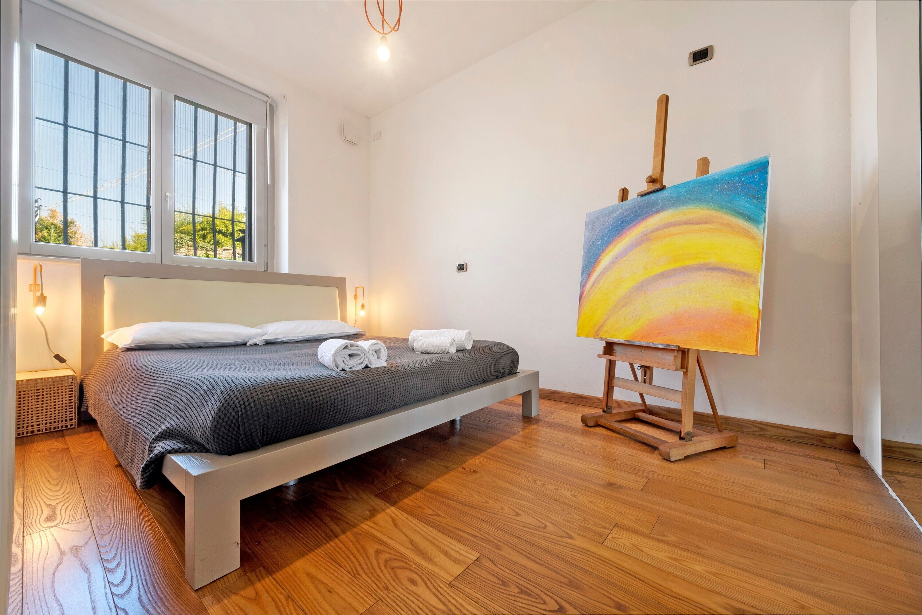 Ein Schlafzimmer mit einem Bett, einem Gemälde auf einem Lehrstuhl und einem Fenster mit Jalousien.