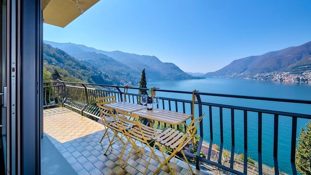 A balcony with a table and chairs, overlooking a lake with mountains in the background. - Dreamy Lakeview Mornings Await! (Orea)