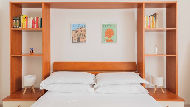 A bedroom with a wooden headboard, two wall-mounted bookshelves, a bed with white bedding, and a small bedside table with a lamp. - Nosdom Properties - National Street (Roma)