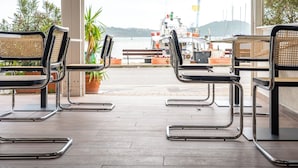 A terrace with metal-framed chairs and a bench, overlooking a marina with boats and a hilly landscape.