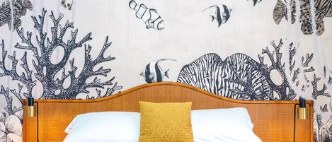Una cama con cabecera de madera, un cojín color mostaza y uno blanco. Hay dos mesitas de noche con lámparas. La pared presenta un papel tapiz con temática marina que muestra peces y coral.