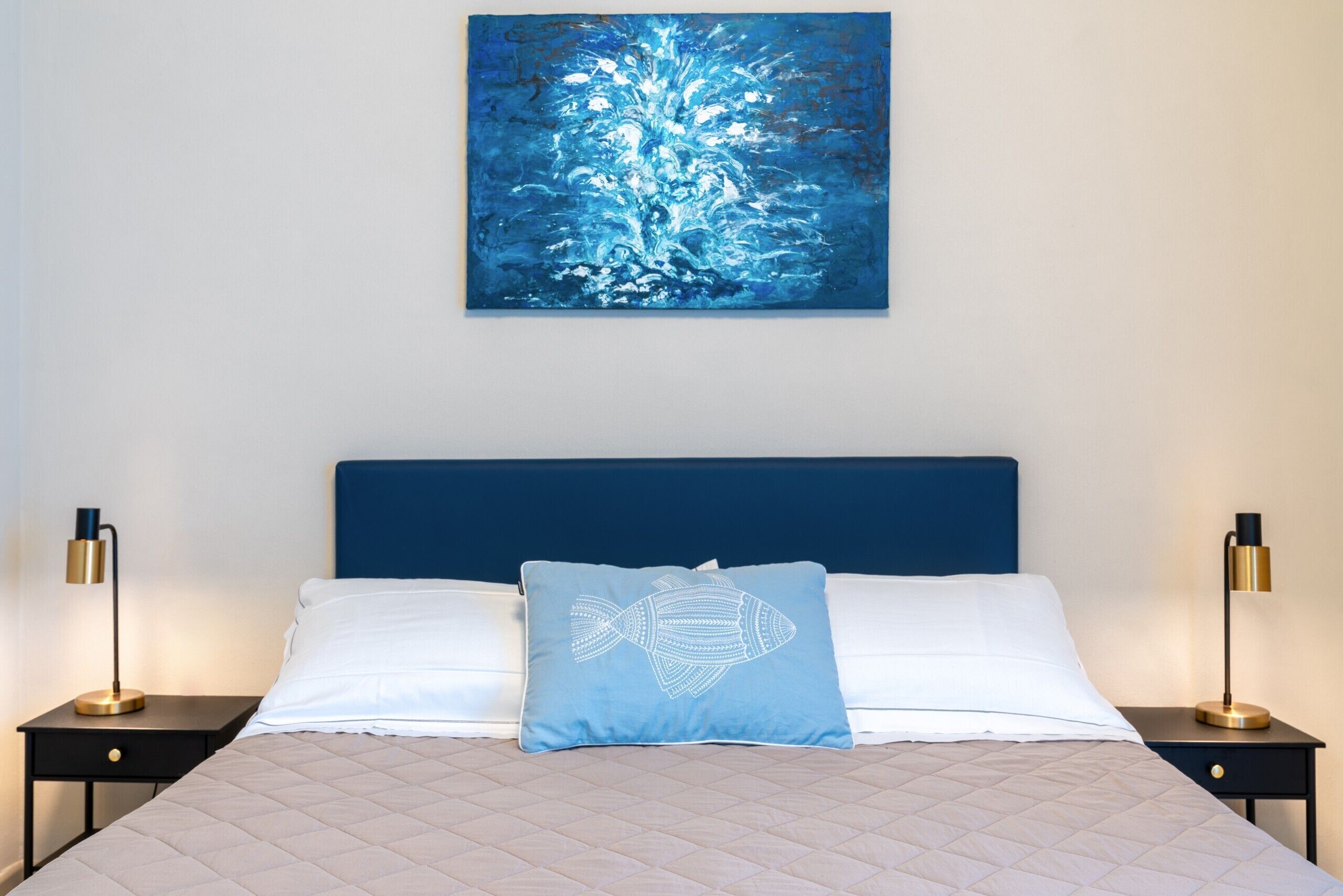 Un lit bien fait, recouvert d’une courtepointe matelassée, un oreiller bleu orné d’un motif de poisson et une tête de lit bleue. De chaque côté du lit, on trouve deux tables de chevet sur lesquelles sont posées des lampes.