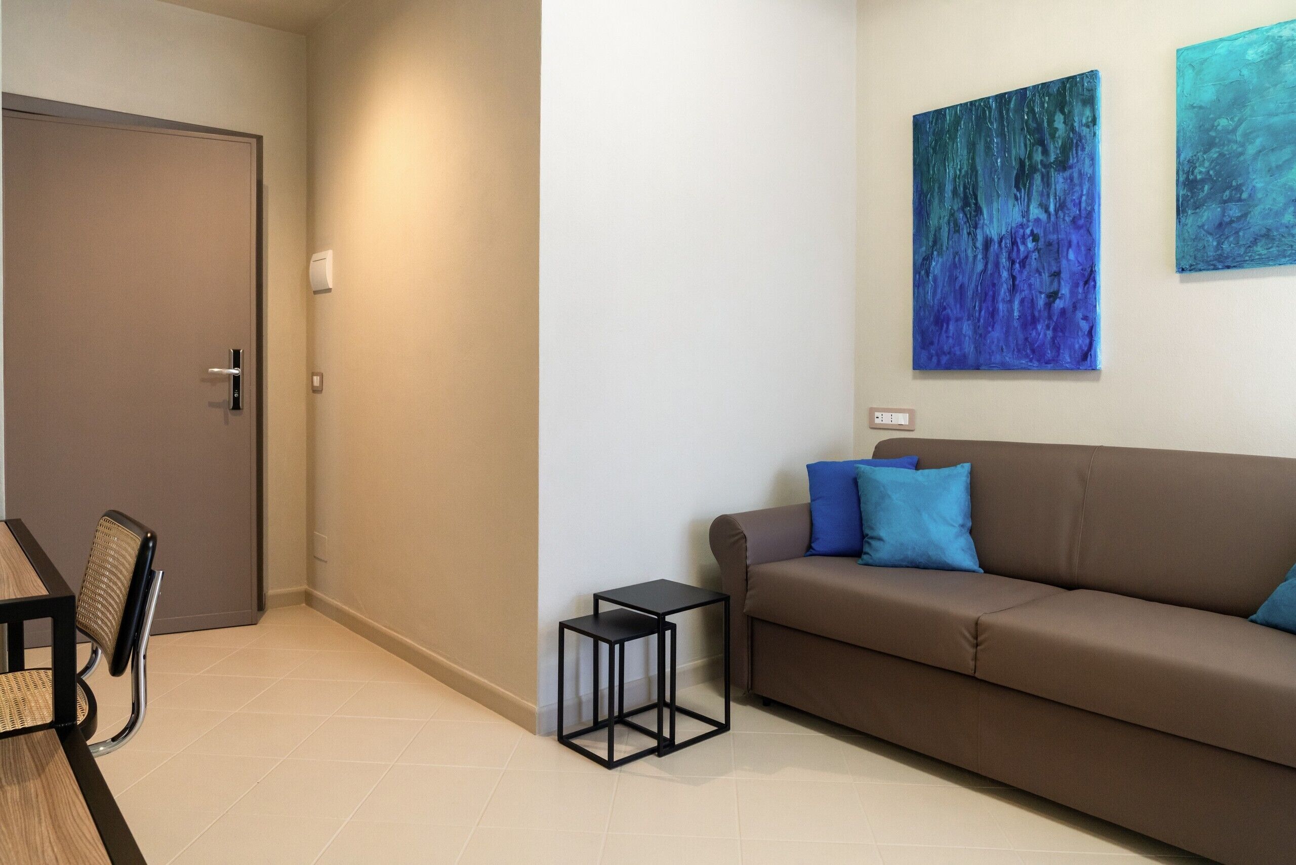 A modern living room with a brown sofa, two blue cushions, a small black side table, a wicker chair, and two abstract paintings on the wall.