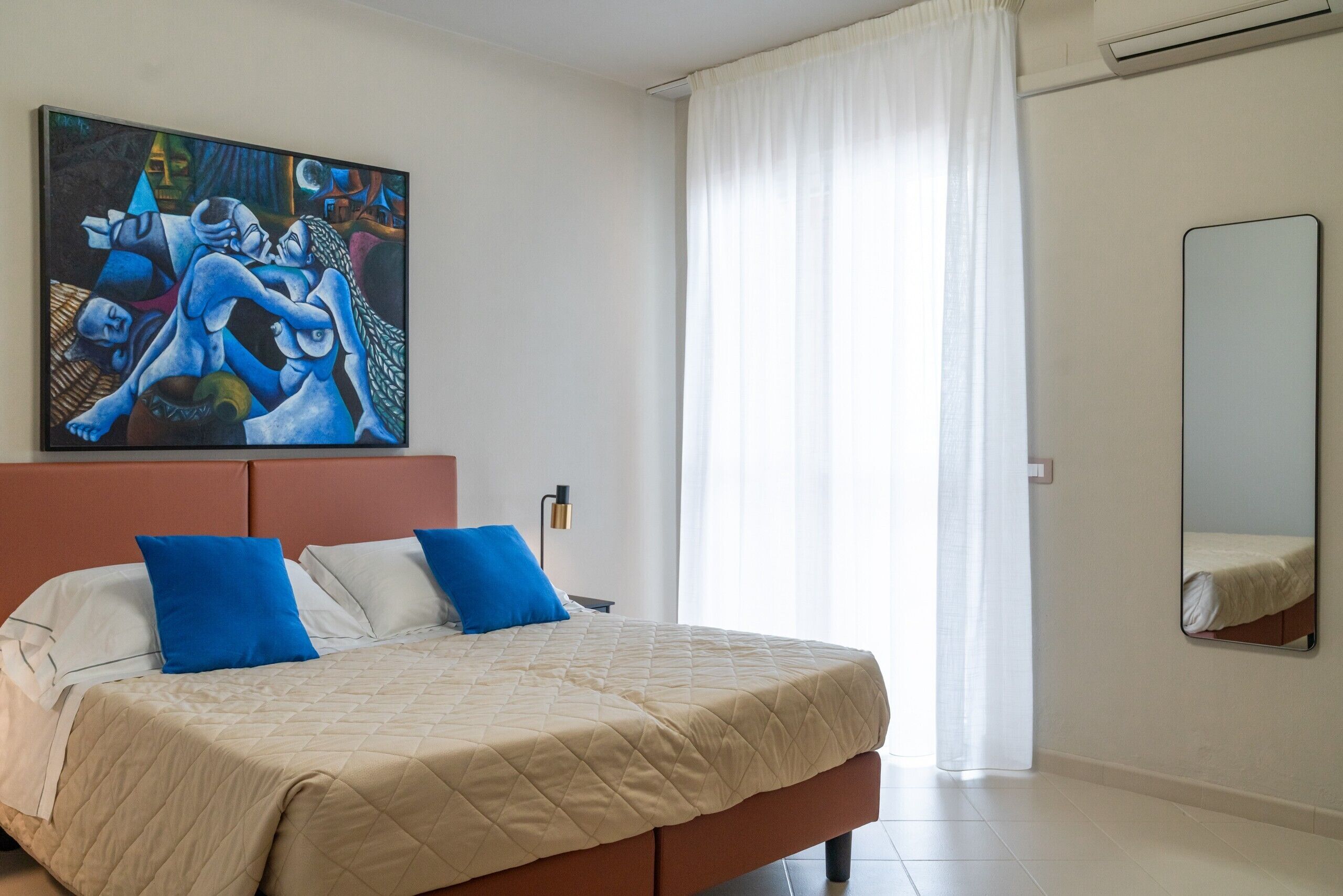 A bedroom with a large bed, a painting on the wall, a mirror, and a window with curtains.