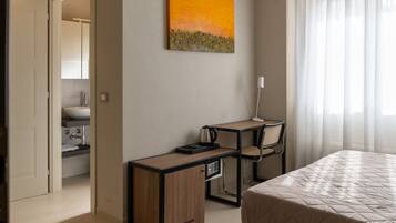 Une chambre d’hôtel avec un lit, un bureau et une chaise, un tableau accroché au mur, et une salle de bain visible à travers une porte ouverte.