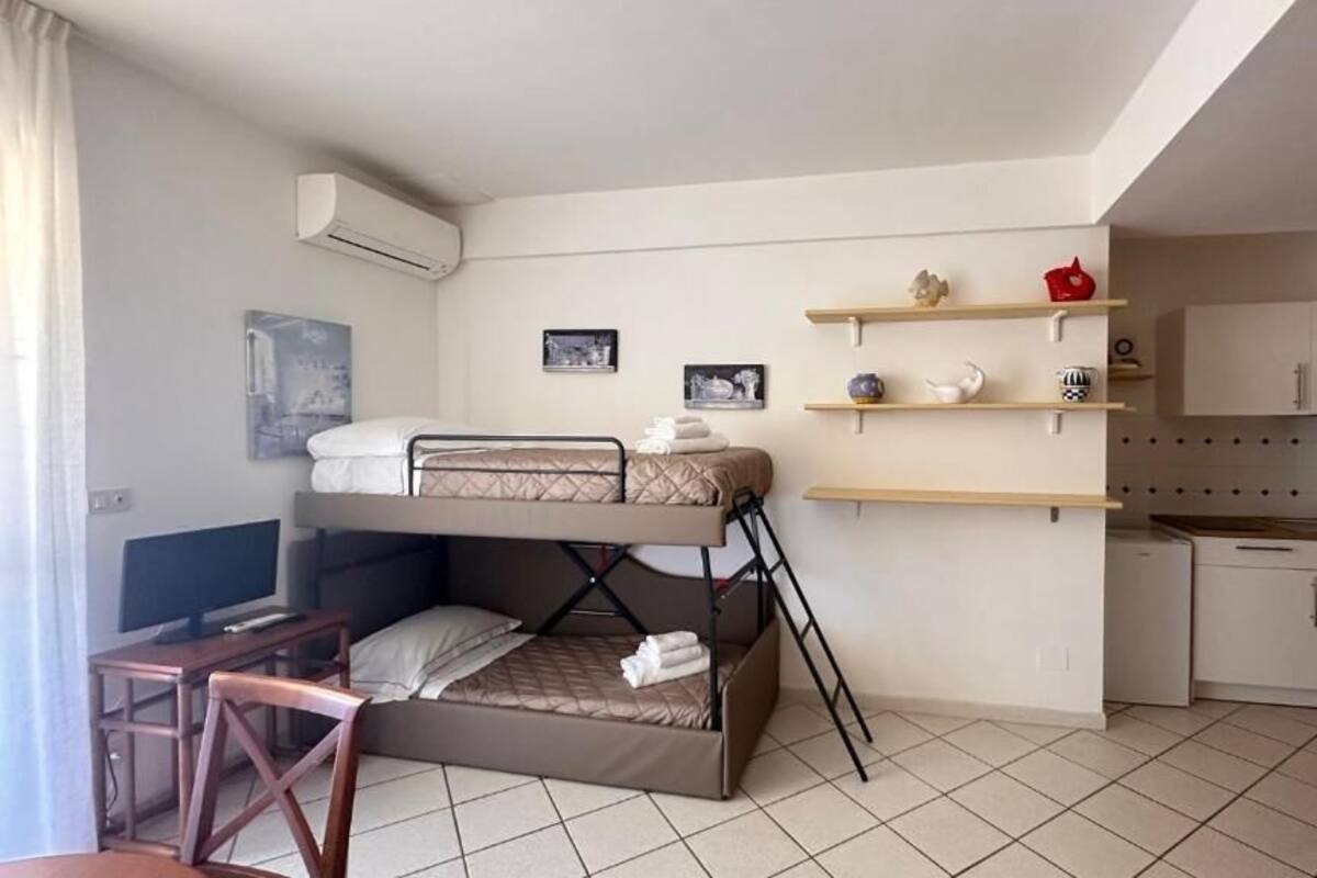 Een compacte hotelkamer met een stapelbed, een klein bureau, een stoel, een televisie, een kitchenette en planken met decoratieve voorwerpen.