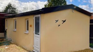 A small, single-story building with a beige exterior, a white door, and three windows. - Modern Private Guest Suite Near Heathrow Free Parking & Garden (Feltham)