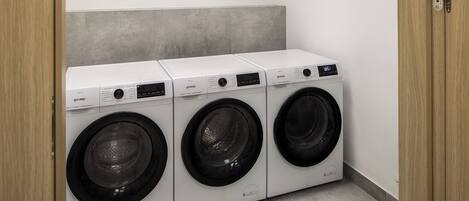 A laundry room with three white front-loading washing machines, a grey tiled wall, and a wooden door.