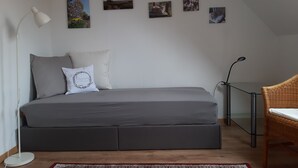 Ett sovrum med en grå bäddsoffa, en vit lampa, en flätad stol och fyra inramade tavlor på väggen.