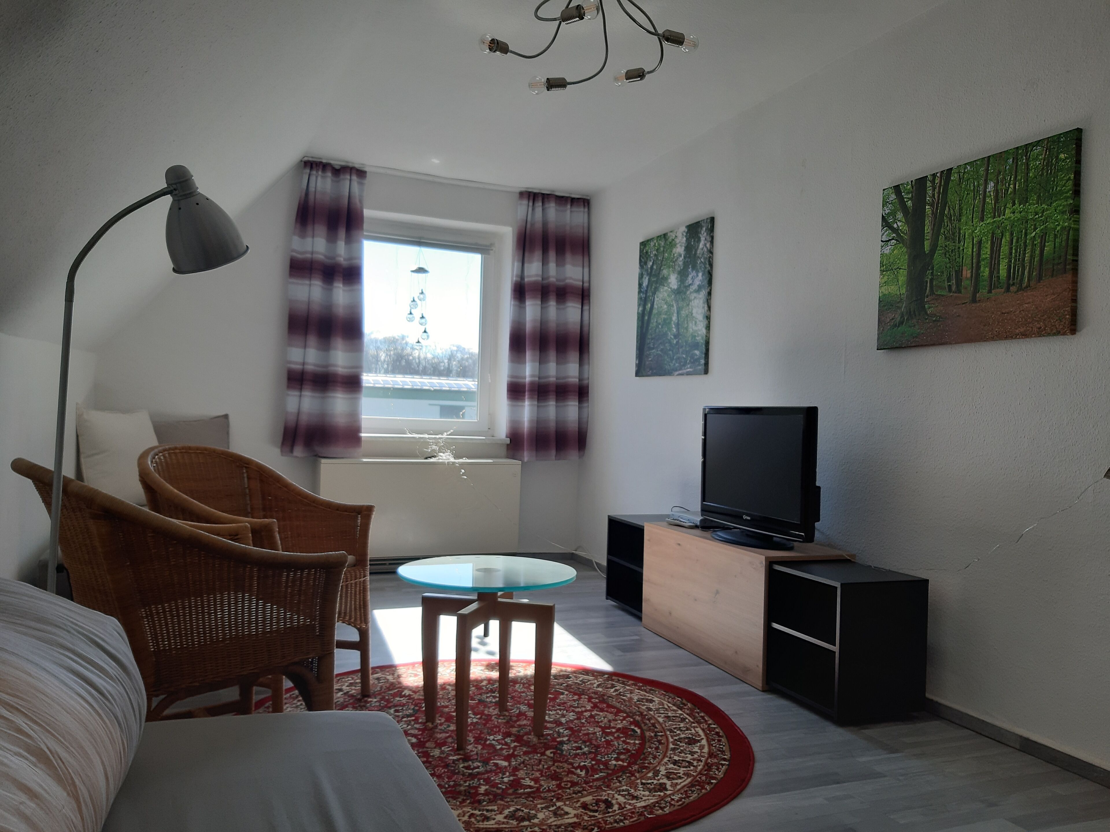 A room with a bed, a chair, a small table, a TV on a wooden cabinet, a red rug, a window with curtains, and a painting on the wall.