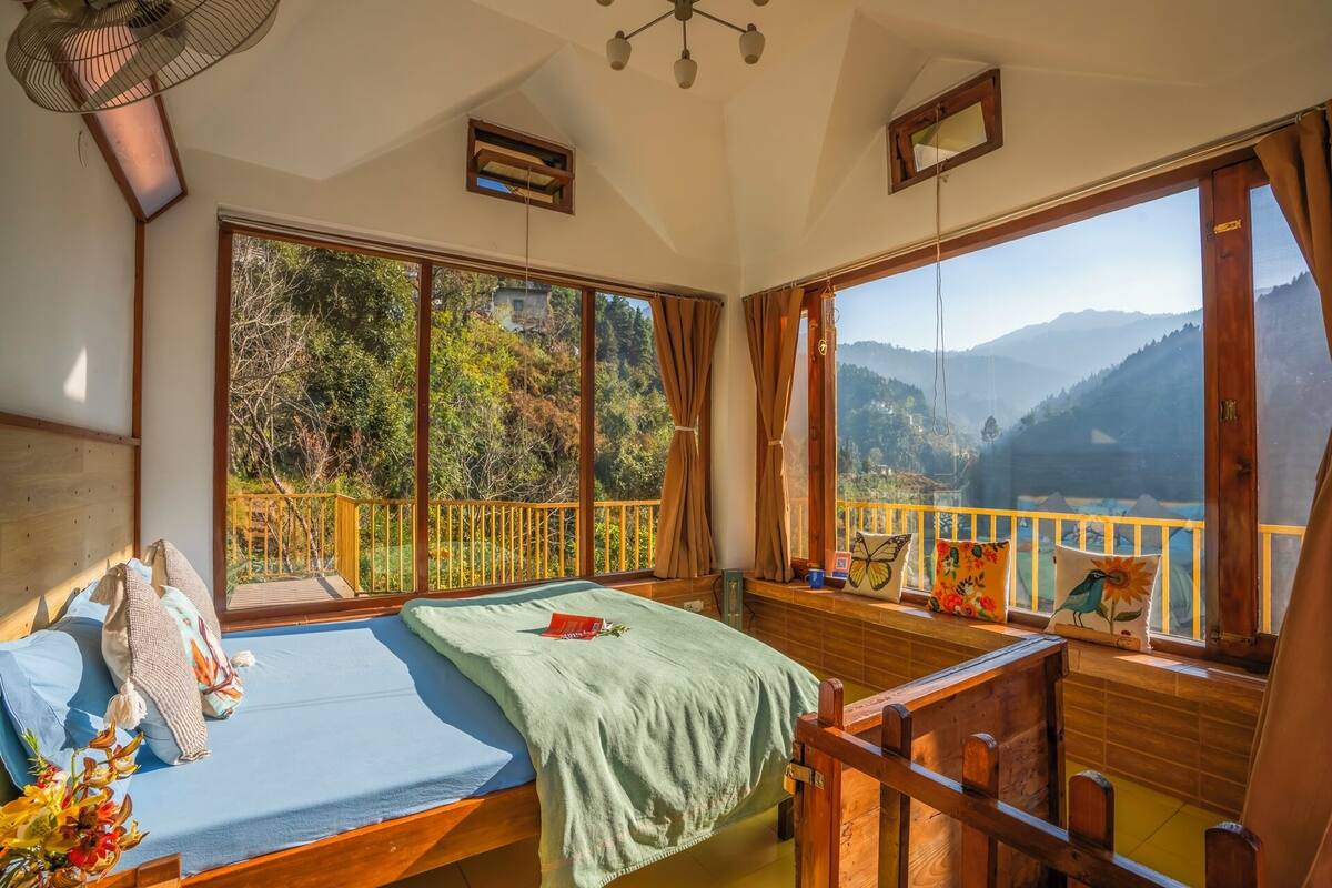 Quarto com cama, uma cadeira de madeira, janela com vista para montanhas e varanda com guarda-corpo.