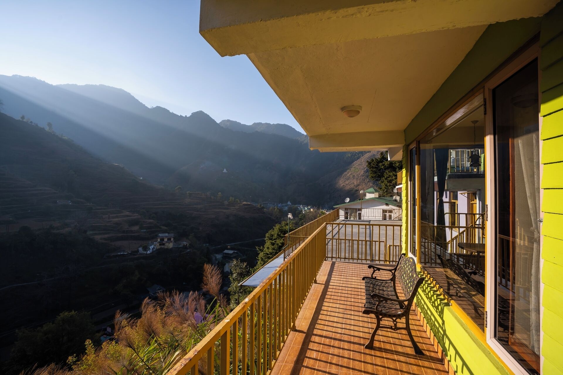 A balcony with a wooden floor and a bench, overlooking a valley with mountains in the distance.