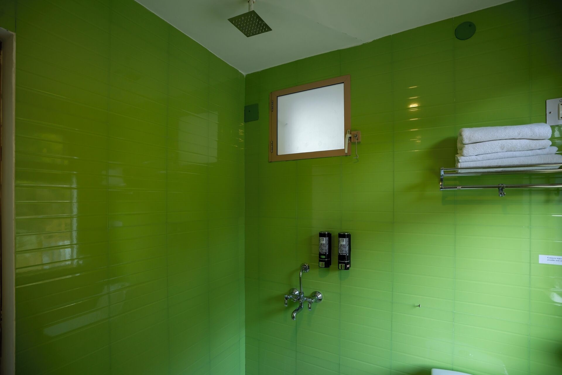 A bathroom with green tiled walls, a white-framed mirror, a towel rack with folded towels, and two black dispensers.