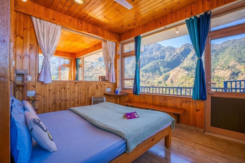 A wooden cabin room with a bed, a bench, and large windows offering a mountain view.