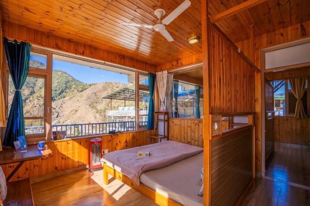 Quarto de cabana de madeira com cama, escrivaninha e janela com vista para uma paisagem montanhosa.