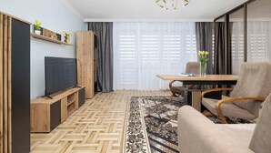 Ein modernes Wohnzimmer mit Fernseher, Holzmöbeln, einem gemusterten Teppich und einem Essbereich mit Tisch und Stühlen.