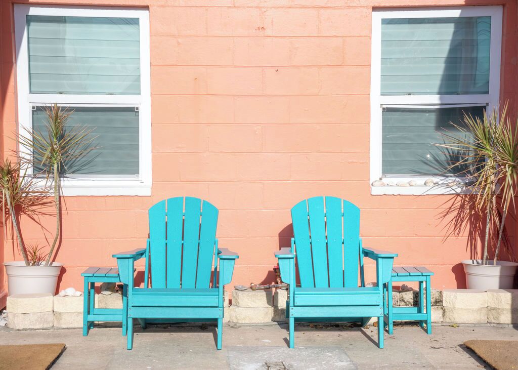Deux chaises Adirondack turquoise sont placées devant un bâtiment doté de deux fenêtres et d’un mur de couleur pêche.