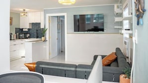Ein modernes Wohnzimmer mit grauem Sofa, einem flachbildfernseher darüber angebracht und einer Küchenzeile mit weißen Schränken und einer blauen Spritzschutzwand.