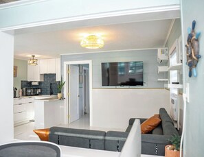 A modern living room with a grey sofa, a flat-screen TV mounted above it, and a kitchen area with white cabinets and a blue backsplash. - Modern & Quiet Oasis, 1 minute walk to the Beach (Madeira Beach)