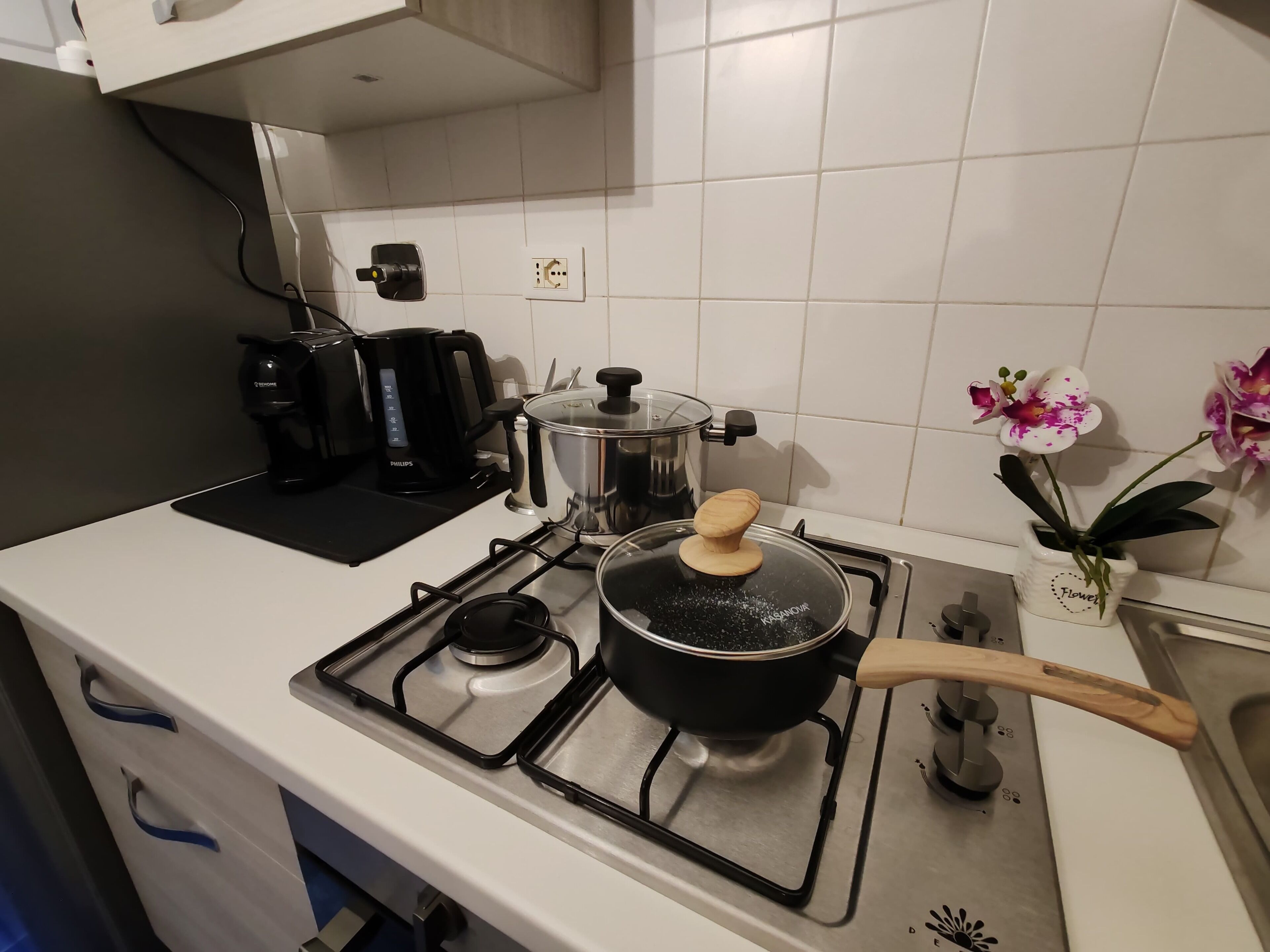 Et køkken med gaskomfur, kaffemaskine, gryde på komfuret og en blomstervase.