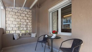 Ein Balkon mit einem gemusterten Sichtschutz, einem runden Tisch mit zwei Stühlen und einem Fenster mit Blick auf ein Gebäude.