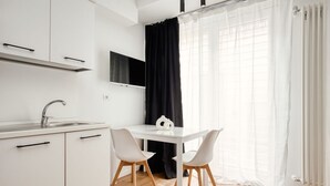 Een moderne keuken met witte kastjes, een spoelbak en een flatscreen televisie boven de aanrecht. Er staat een eettafel met twee stoelen, en een zwart gordijn is gedeeltelijk opengetrokken.