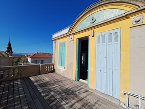 A balcony with a wooden floor, a yellow wall with a blue door, and a view of the cityscape in the background. - Pointe Rouge à Marseille (Marseille)