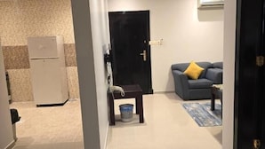 A modern living room with a sofa, a coffee table, and a rug. There is a door leading to another room and a refrigerator in the background. - Makarem Najd 5 - Al Aqiq (Riyadh)