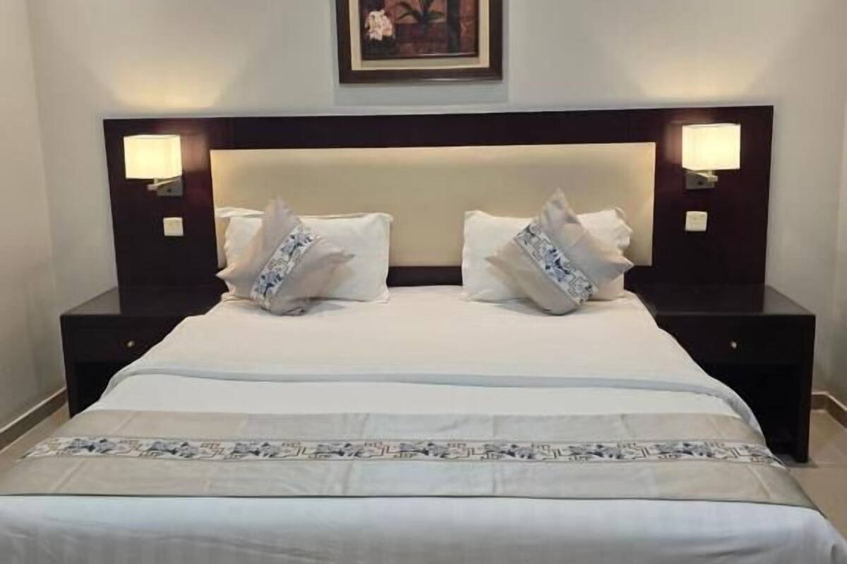 Een hotelkamer met een groot bed, twee nachtkastjes met lampen, een lijst met een afbeelding boven het bed, en een decoratieve loper op het bed.