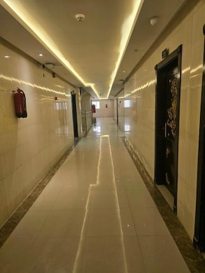Un pasillo largo e iluminado, con un extintor a la izquierda, una puerta con panel decorativo a la derecha y un piso de baldosas impecables.