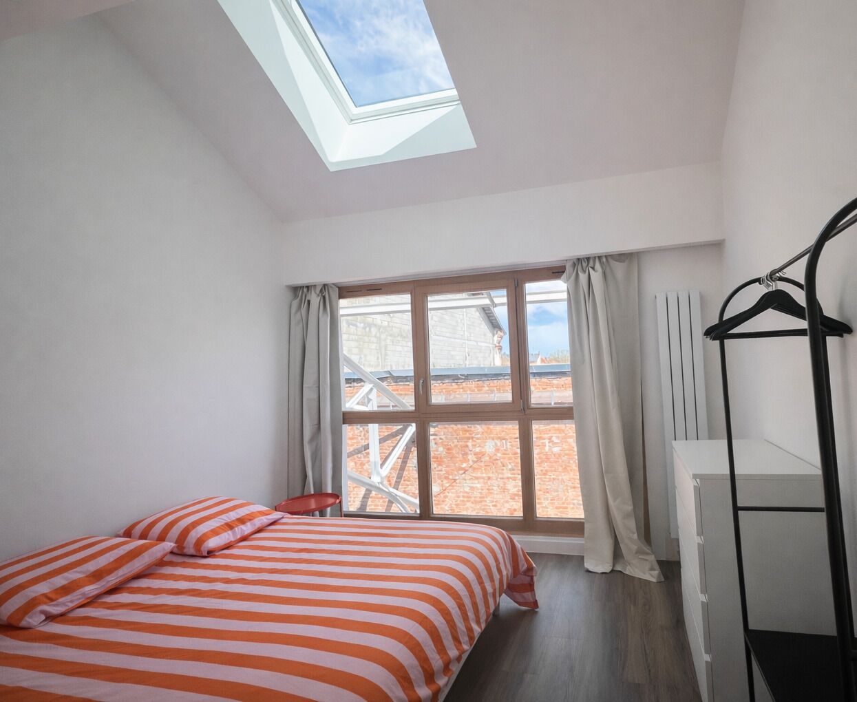Ein Schlafzimmer mit Dachfenster, einem Bett mit gestreifter Bettwäsche, einem Heizkörper und einem schwarzen Kleiderständer.