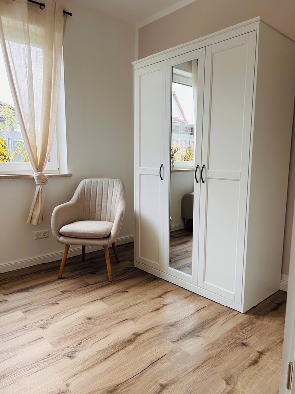 A room with a wooden floor, a white wardrobe, a chair, and a window with sheer curtains. - Vacation home Lounis - Apartment 1 (Bützow)