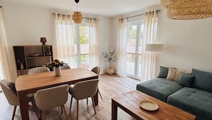 A spacious living area with a wooden dining table, chairs, a sofa, and a coffee table. There is a kitchen area with a stove and cabinets, and a large window providing natural light. - Vacation home Lounis - Apartment 1 (Bützow)