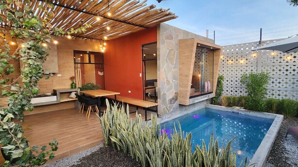 Een modern buitenzwembad met een houten terras, zitplaatsen en een stenen muur met een geometrisch patroon.