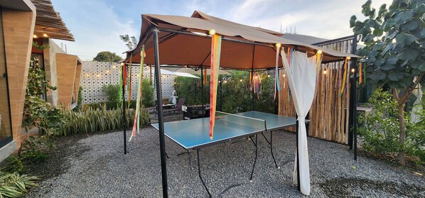 Une table de ping-pong installée à l’extérieur, sous un auvent, sur un sol de gravier, entourée de plantes et d’une clôture en bois.