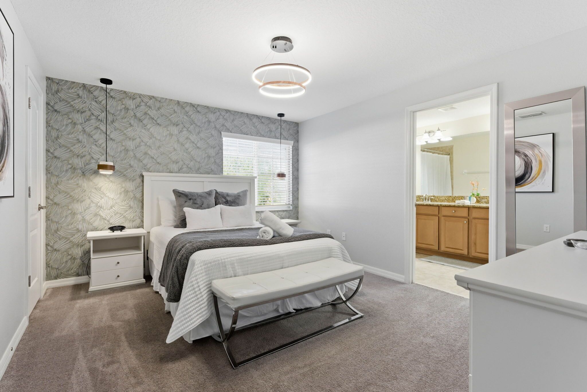 A bedroom with a bed, bedside tables, a bench, a window with blinds, and a bathroom visible through an open door.