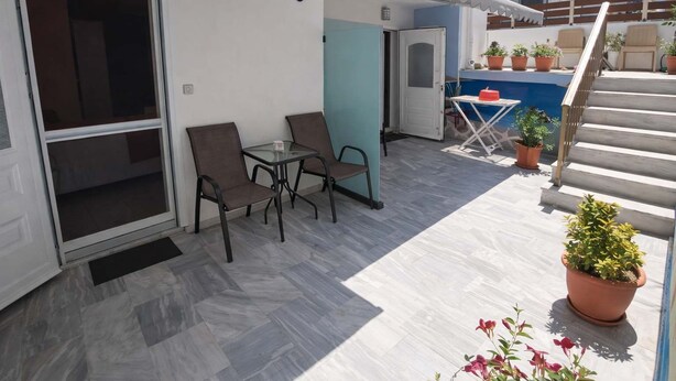 Eine Terrassenfläche mit einem Glastisch und Stühlen, einem kleinen Tisch mit einem roten Gegenstand und Topfpflanzen.