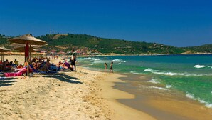Ein Strand mit badenden und sonnenden Menschen, klares blaues Wasser und eine hügelige Landschaft im Hintergrund.