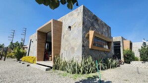 Un bâtiment moderne avec une façade en pierre, une porte d’entrée en bois et un petit jardin agrémenté de cactus et de fleurs jaunes.