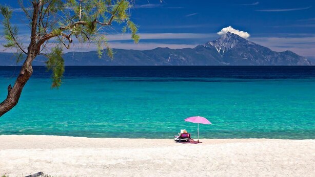 Ein Strand mit klarem türkisblauem Wasser, ein Berg im Hintergrund und eine Person unter einem rosa Sonnenschirm.
