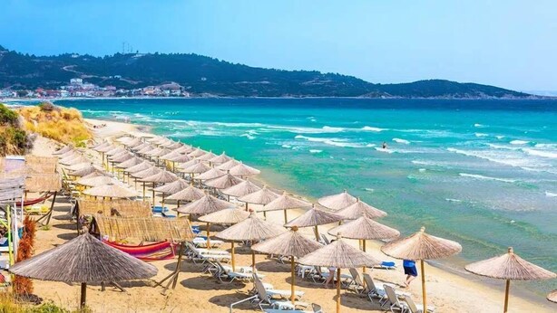 A beach with rows of sun loungers and thatched umbrellas, a clear blue sea, and a distant town on the hillside. - House Blerina - Beachfront Double Sarti Getaway (Sithonia)
