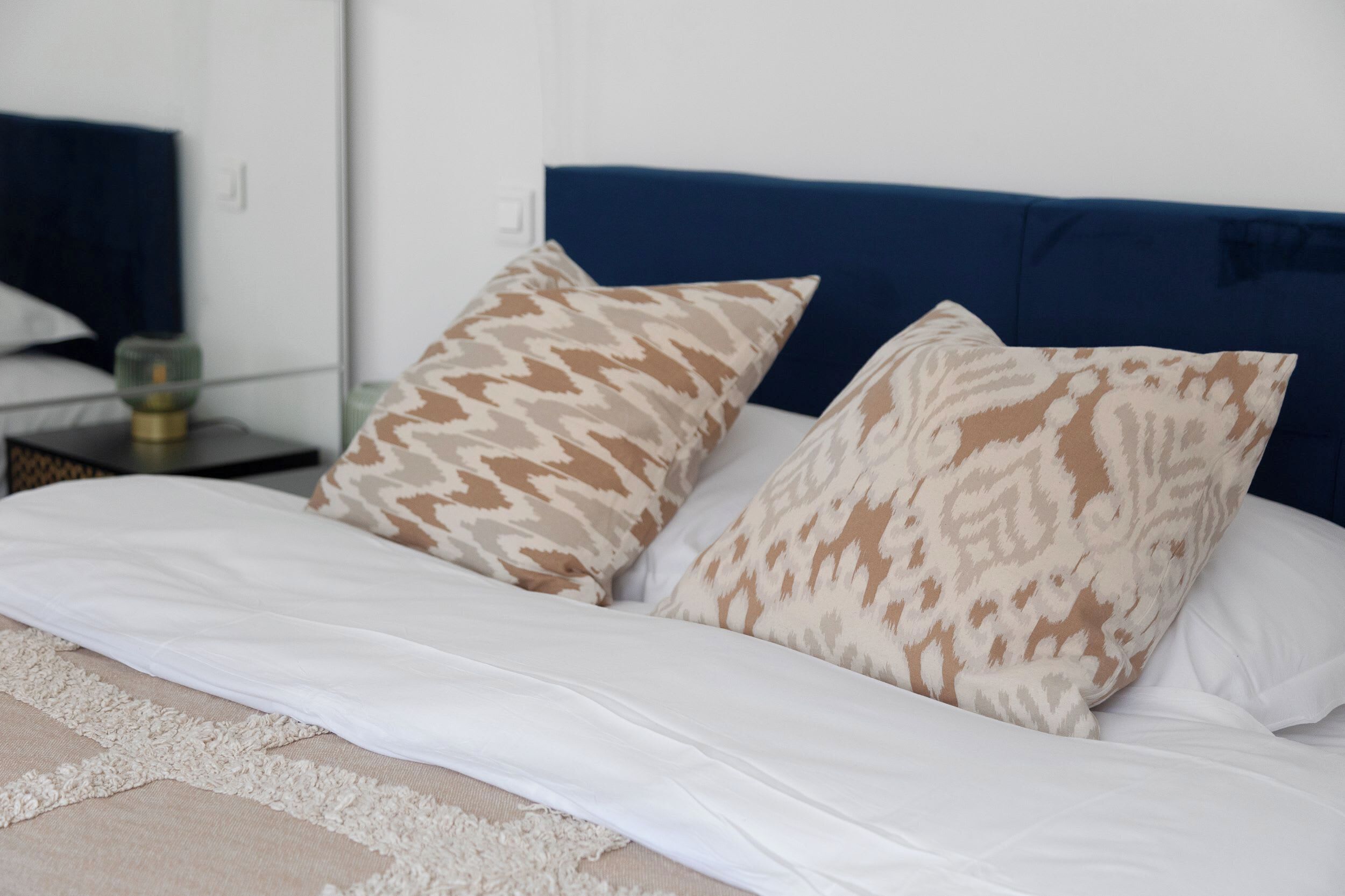 Una cama con ropa de cama blanca y dos almohadas estampadas, un cabecero azul y una mesita de noche con una lámpara.