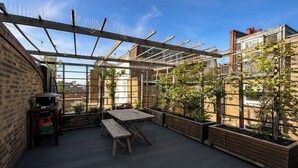 A rooftop patio with a wooden table and benches, surrounded by planters and a pergola with climbing plants. - 1 Bed Apartment - Sleeps 3 - Central London (London)