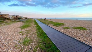 Un sentier qui mène à une plage de galets et d’herbe, avec des bâtiments en arrière-plan.
