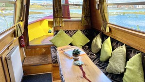 Un intérieur chaleureux avec un banc en bois, des coussins, une petite table agrémentée d’une plante, et une vue sur un plan d’eau à l’extérieur.