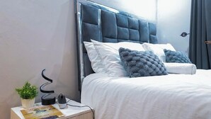 Una cama bien hecha con cabecera capitonada, ropa de cama blanca y dos almohadas decorativas. Se ve una mesita de noche con una lámpara, una planta y un centro de carga.