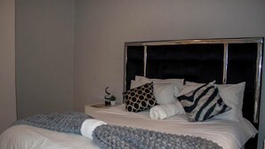 Un lit bien fait, avec une couverture grise et un plaid bleu, une tête de lit noire et une petite table de chevet agrémentée d’une plante.