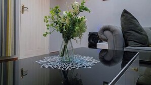 Une pièce avec une table en verre, un vase rempli de fleurs, un chemin de table blanc, un canapé gris et un tableau encadré accroché au mur.